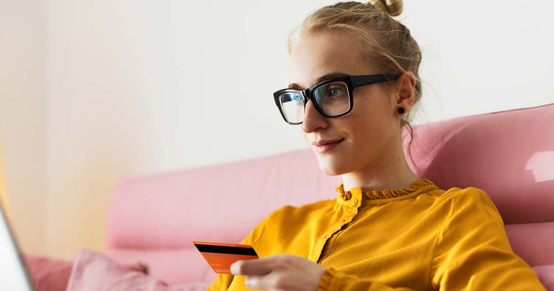 Pige i briller og gul trøje, der sidder i en pink sofa med betalingskort og bærbar computer