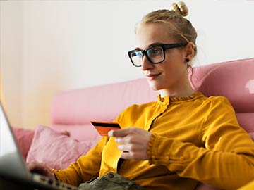 Pige i briller og gul trøje, der sidder i en pink sofa med betalingskort og bærbar computer
